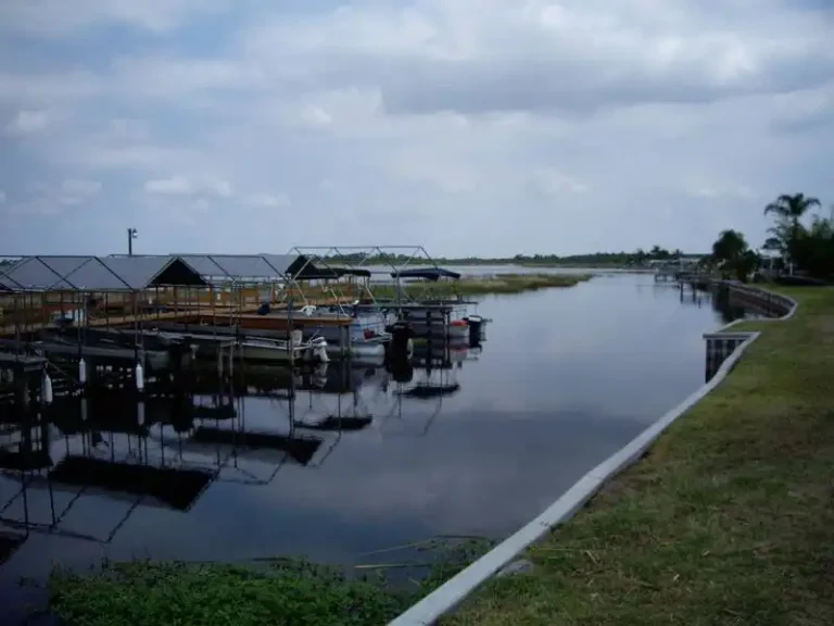 A calm, narrow river winds through Lakeside Ranch, with several covered docks and boats resting under a cloudy sky. Grassy banks line the waterway, offering a serene haven perfect for those seeking tranquility in a 55+ community.