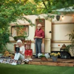 An elderly couple enjoys a picnic outside their RV at Lakeside Ranch, surrounded by greenery and string lights. The woman is seated on a blanket with a tablet, while the man stands holding plates. This peaceful 55+ community offers the perfect backdrop for leisurely moments together.