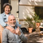 An older couple beams with joy outdoors at Lakeside Ranch, their caravan nestled in a lush garden of plants and outdoor furniture, epitomizing the vibrant life of a 55+ community.