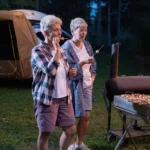 At Lakeside Ranch, a vibrant 55+ community, two people enjoy roasting marshmallows near a grill at their campsite. In the background, a tent is snugly set up, completing this picturesque outdoor escape.