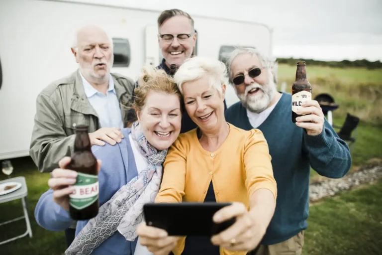 A group of five adults stands outdoors at Lakeside Ranch, smiling and taking a selfie while holding beer bottles. A caravan is visible in the background, capturing the spirit of this vibrant 55+ community.