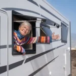 An older woman in a colorful sweater leans out of an RV window, smiling and gesturing with her arm. The landscape features the stunning Lakeside Ranch, nestled in the mountains—a perfect getaway spot for the lively 55+ community.