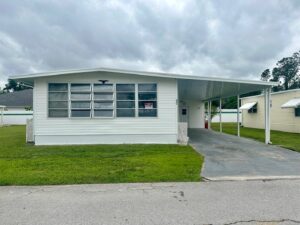 A white manufactured home with large windows and a carport sits gracefully on a well-maintained lawn under a cloudy sky, evoking the simplicity of ranch living.
