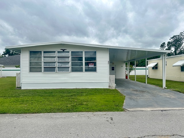 A white manufactured home with large windows and a carport sits gracefully on a well-maintained lawn under a cloudy sky, evoking the simplicity of ranch living.
