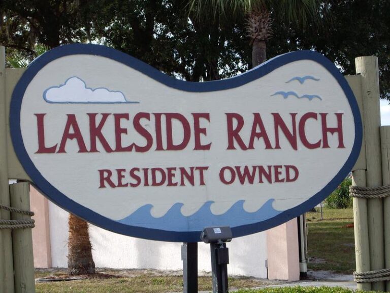 A sign reading "Lakeside Ranch Resident Owned," adorned with decorative clouds and waves, welcomes visitors amid trees and a grassy area. Discover exciting lakeside ranch activities in this serene setting.
