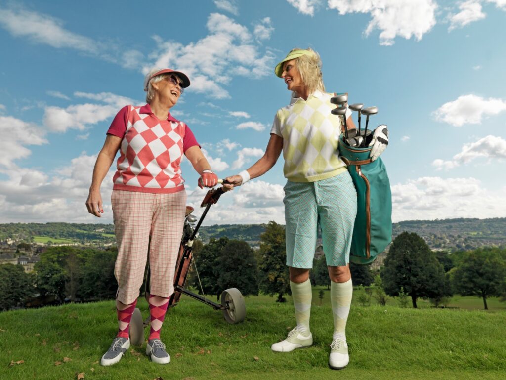 Two women in patterned golf attire smile and chat on a grassy hill with a golf cart. One holds a golf bag under the blue sky with clouds, enjoying the lakeside ranch activities as part of their park adventures.