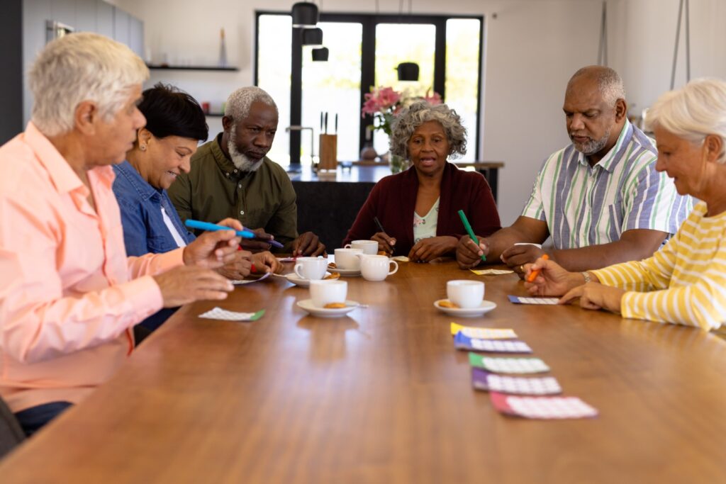 In a well-lit room reminiscent of park activities, a group of older adults sits at a table, holding pens, with bingo cards and cups of tea in front of them.