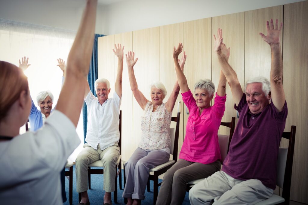 A group of elderly people sitting in chairs, raising their arms in a light and airy room, engage in lakeside ranch activities guided by a person at the front.