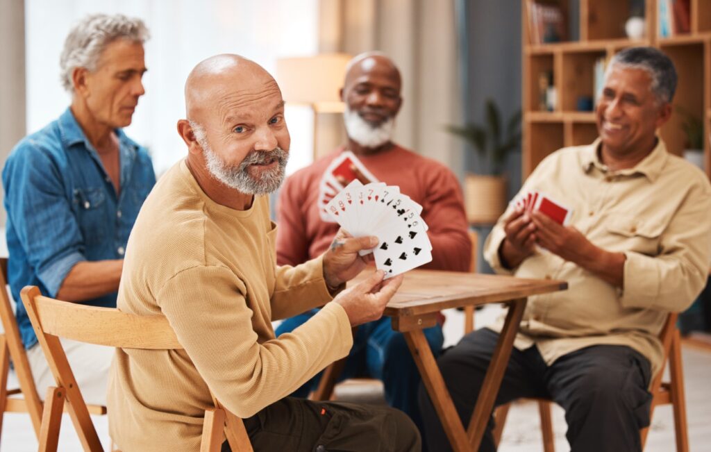 Four men are sitting around a table playing cards, enjoying the camaraderie of their lakeside ranch activities. The man in the foreground is smiling and holding a fan of cards, clearly relishing this leisurely afternoon by the water.