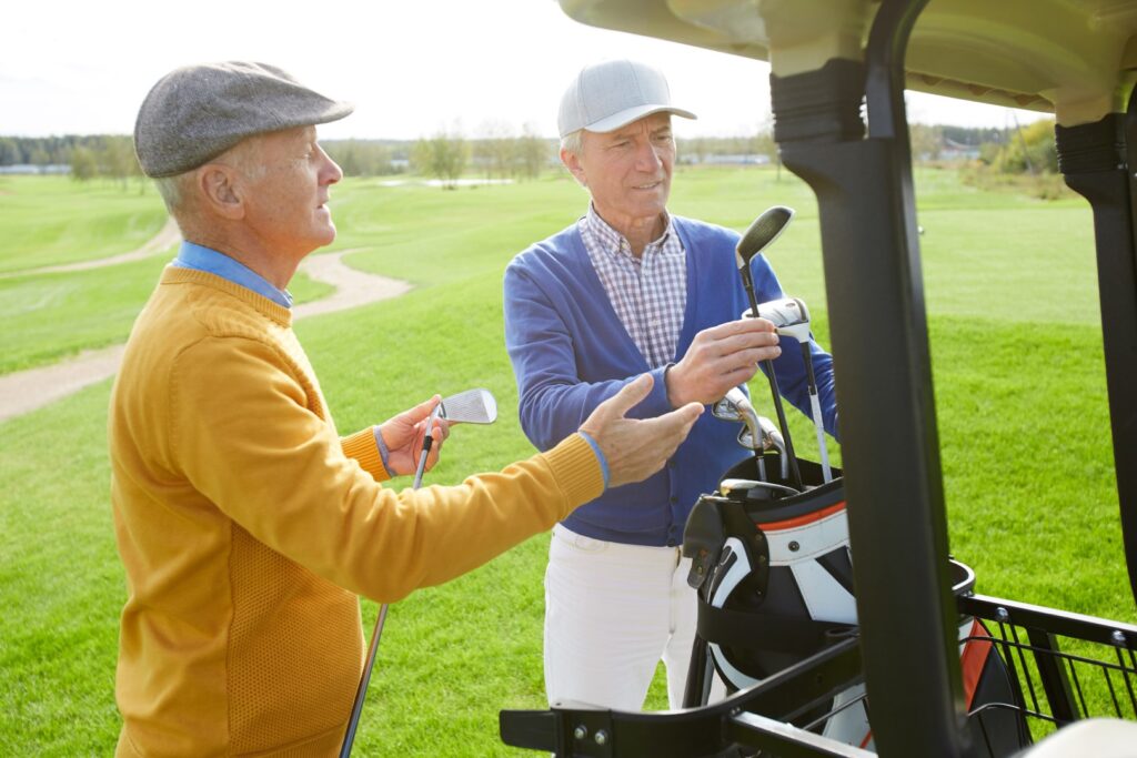 Two older men stand by a golf cart on a sunny day, discussing park activities while holding golf clubs on a lush green golf course.