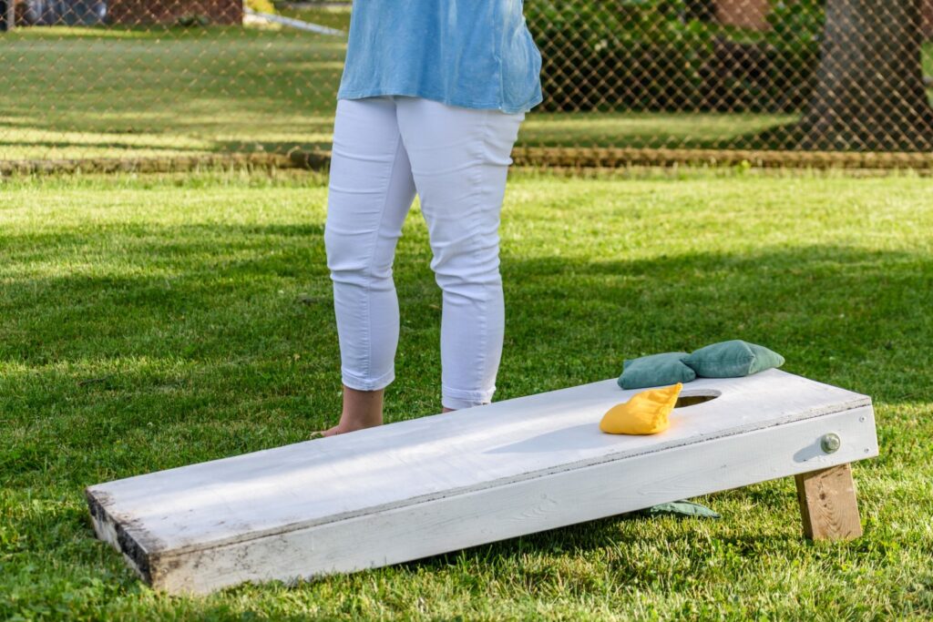 A person stands next to a cornhole board with blue and yellow bean bags on it in a grassy yard, enjoying one of the popular lakeside ranch activities.
