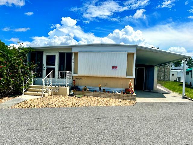 A white mobile home with a covered carport, front steps, and a small landscaped area, set against a bright blue sky with clouds.